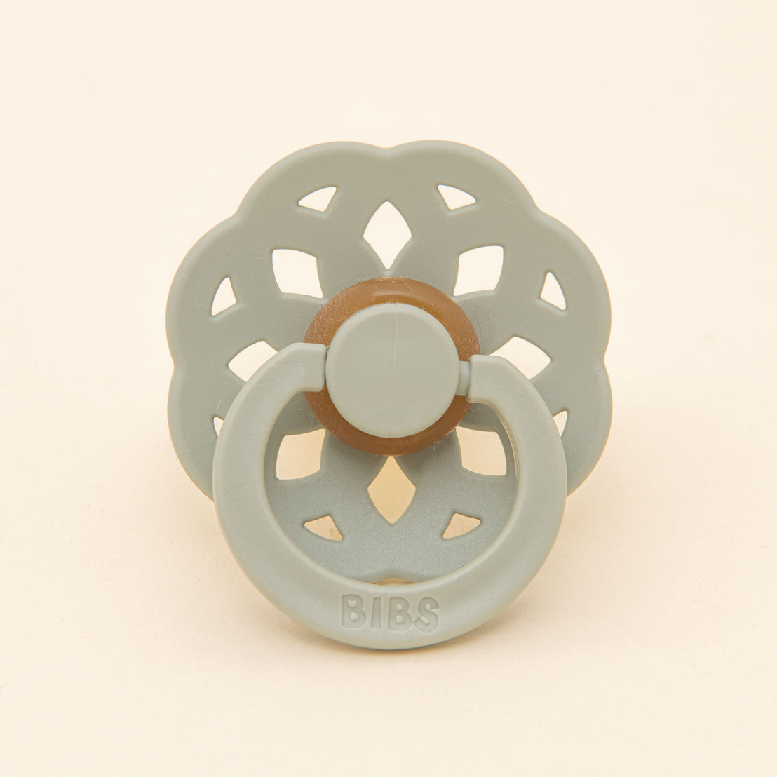 The sage green Mila Pacifier with a round silicone nipple and a flower-shaped shield set against a soft beige background.
