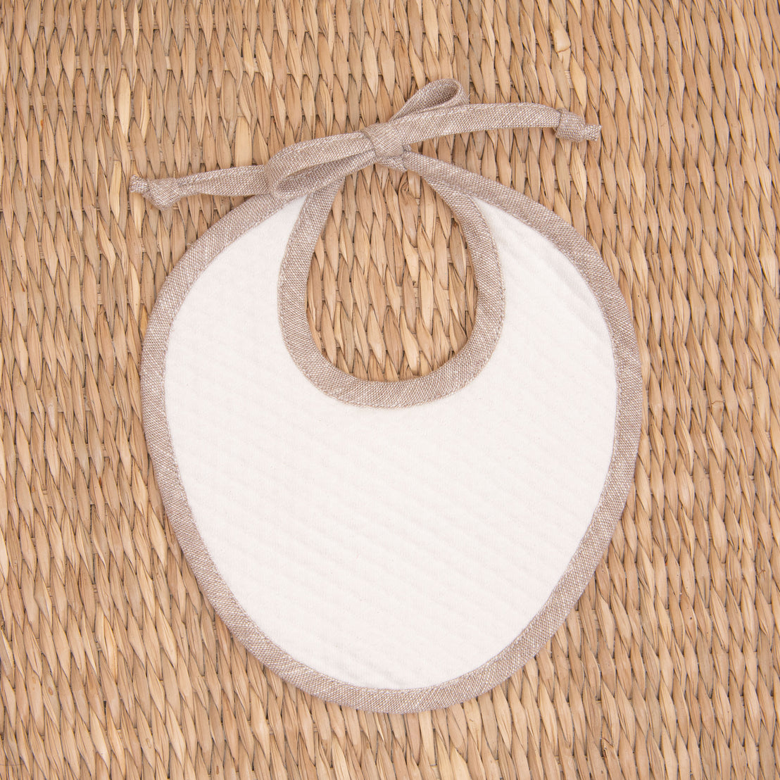 Flat lay photo of the tan Silas Linen Trim Bib. The bib is made from textured cotton in light ivory