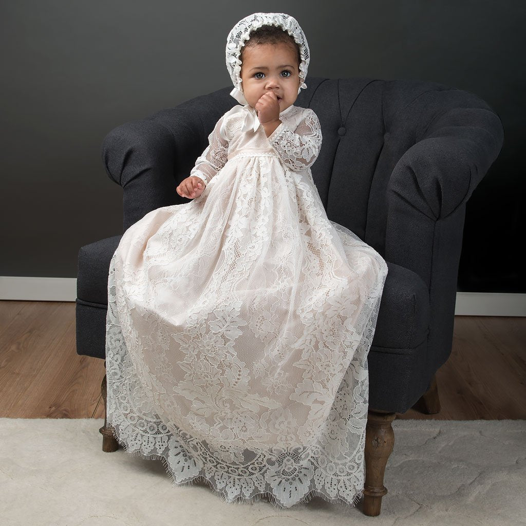 Baby girl on chair with hand over mouth, wearing the Long Sleeve Victoria Christening Gown. Pictured on a dark chair.