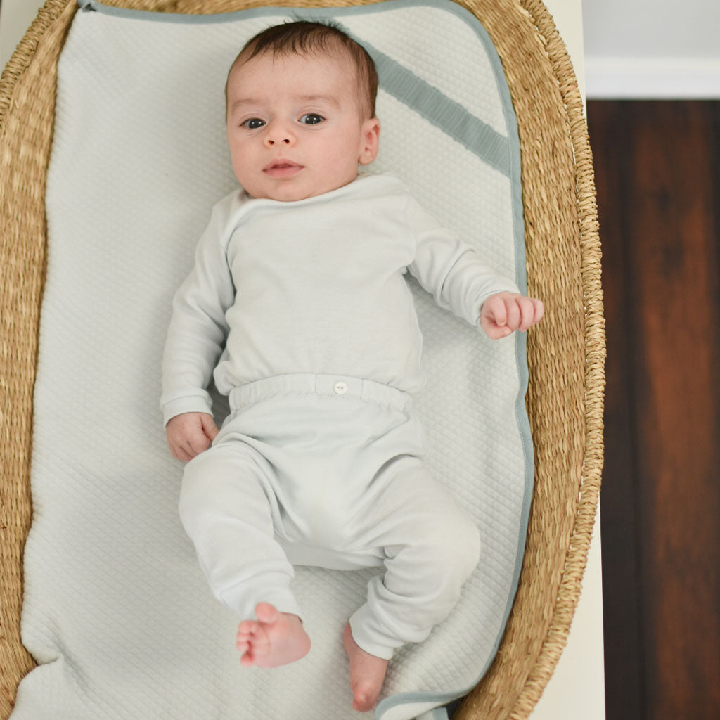 A baby wearing the Aiden Pima Top & Leggings onesie lies comfortably in a wicker bassinet, looking upwards with a gentle gaze.