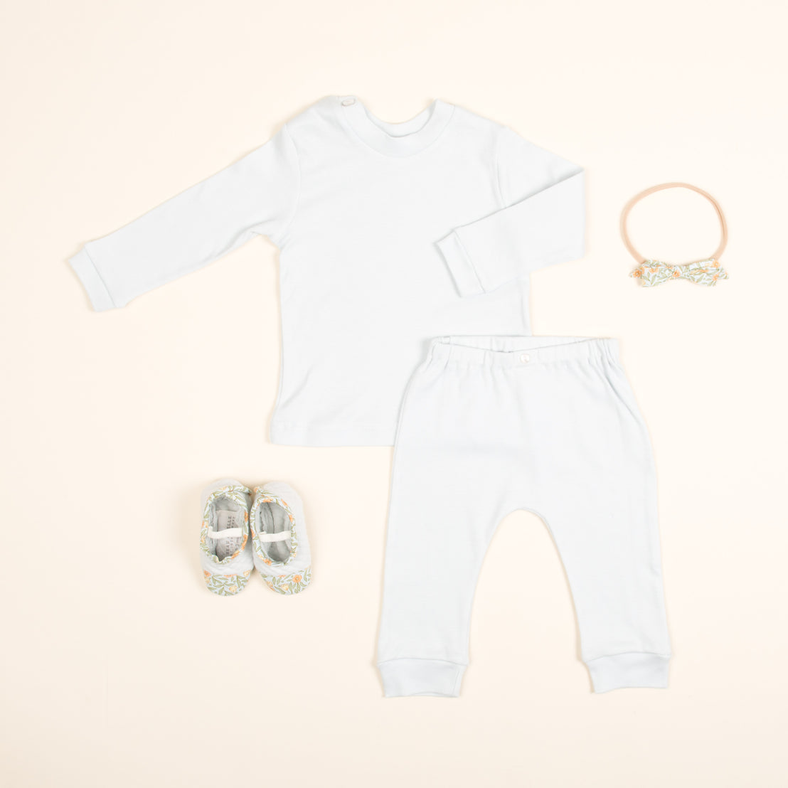 A flat lay of an Aiden Pima Top & Leggings baby outfit on a beige background, featuring a white long-sleeve shirt, light grey pants, matching floral shoes, and a floral headband.