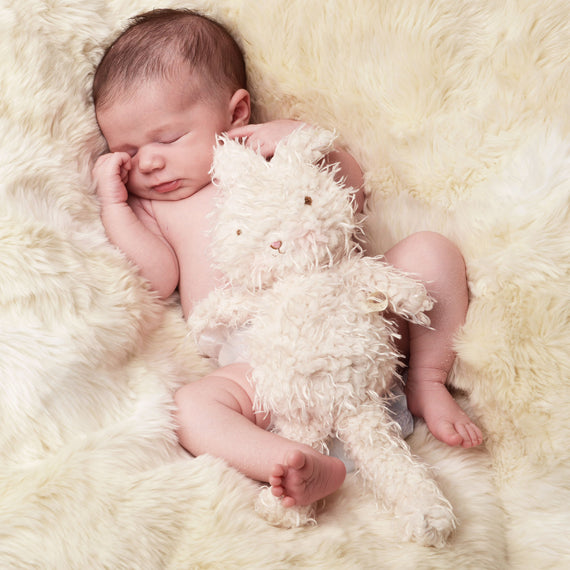 A newborn baby sleeps peacefully on a cream-colored fluffy blanket, clutching a soft, plush white Shaggy Hoppy Bunny toy, dressed in vintage baby clothes newborn layette.