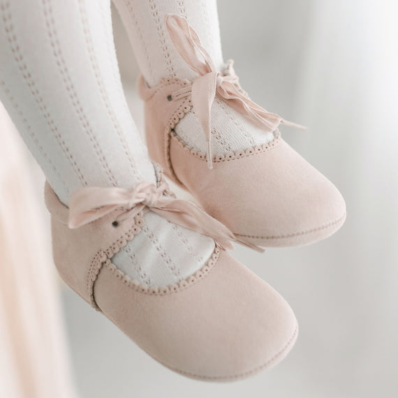 Pair of Elizabeth Suede Tie Mary Janes in pink for christening, featuring soft texture and intricate stitching, gently hanging against a blurred background.