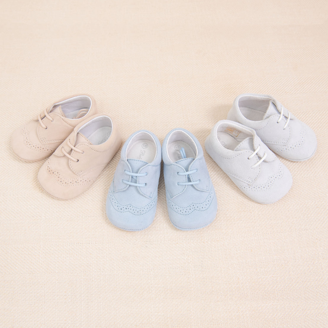 Three pairs of Baby Beau & Belle Boys Suede Shoes, two beige and one blue, arranged in a row on a beige textured background.