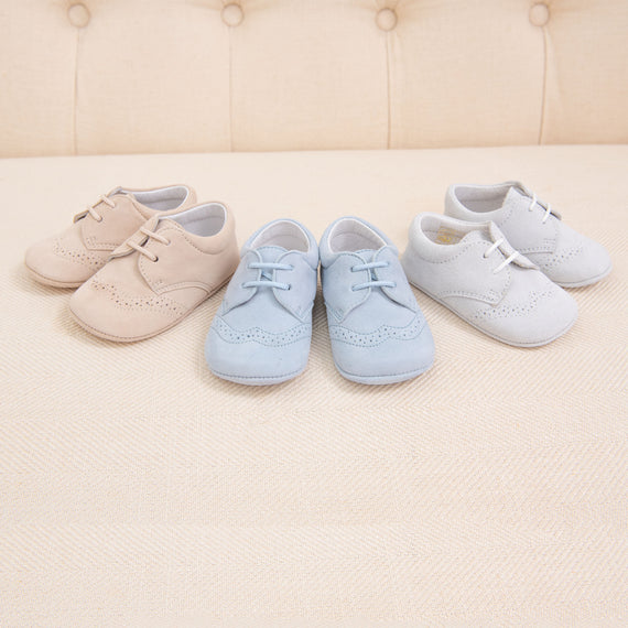 Three pairs of Baby Beau & Belle Boys Suede Shoes in beige, blue, and white lined on a beige textured surface with a cushioned backrest in the background.