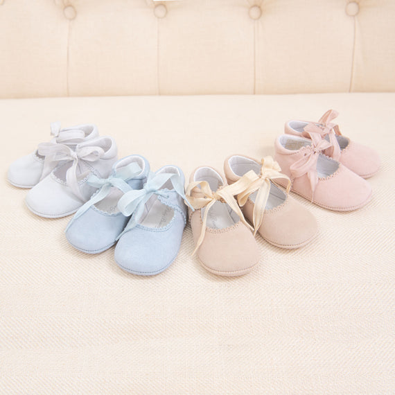 Three pairs of Girls Suede Tie Mary Janes in blue, beige, and pink, each tied with a ribbon, arranged in a row on a cream background with a tufted cushion behind.