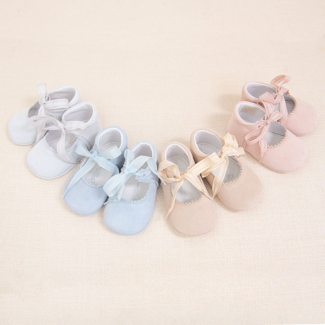 Four pairs of Girls Suede Tie Mary Janes—two pink and two blue—arranged on a beige fabric background, each tied with matching ribbon laces.
