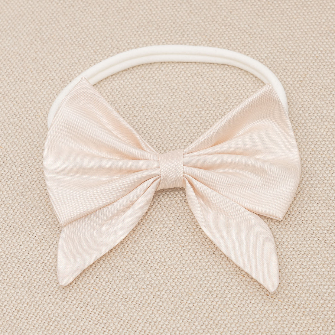 A Elizabeth Bow Headband displayed on a textured beige background, ideal for upscale or vintage baptism events. The bow is neatly tied and centrally positioned on the headband.