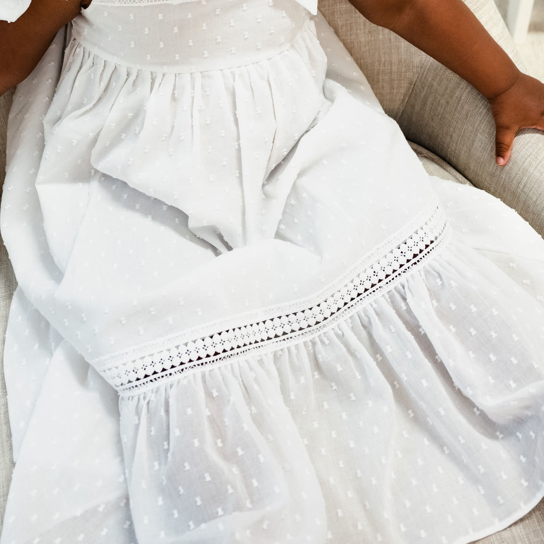 A close-up of a toddler sitting on a sofa, wearing the Mila Cotton Gown. The photo showcases the dotted patterns and lace details, as well as, the texture and design of the gown.