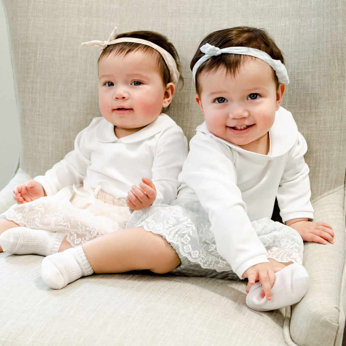 Two cheerful baby girls, dressed in the Isla Bubble Skirt Set with heirloom lace skirts and matching Isla Tie Headbands, sitting together on a light gray sofa.