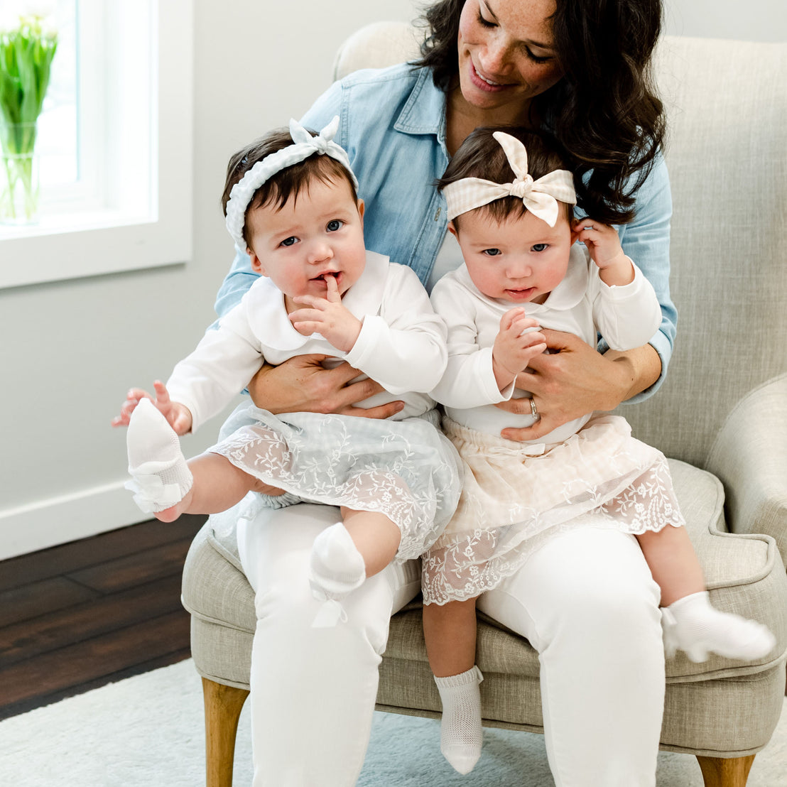 A mother sitting in a chair holding her newborn twin girls, dressed in matching Isla Bubble Skirt Sets with white onesies and matching Isla Tie Headbands. The room has a bright and cozy vintage interior.