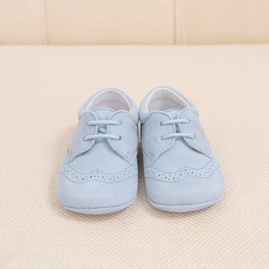 A pair of Baby Beau & Belle boys suede shoes in baby blue with laces, featuring classic brogue detailing, displayed on a soft beige fabric background.
