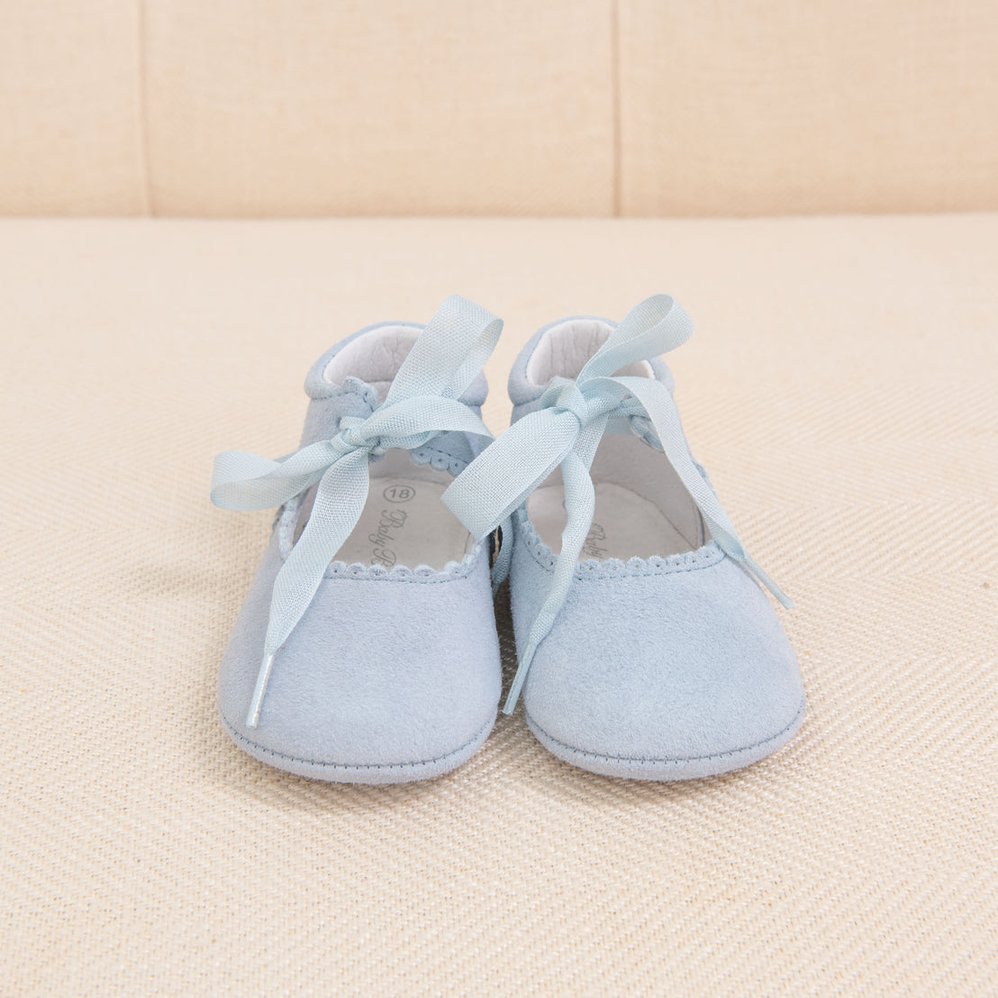 A pair of Girls Suede Tie Mary Janes handmade shoes with light blue ribbons and scalloped edges, sitting on a textured beige fabric background.