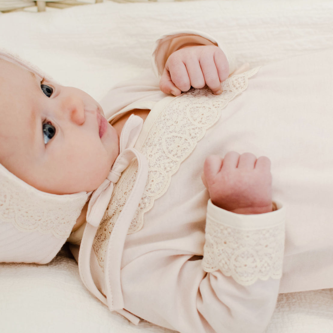 An infant with blue eyes, wearing an Evelyn Knot Gown with lace details, lying on a white bed and looking to the side.