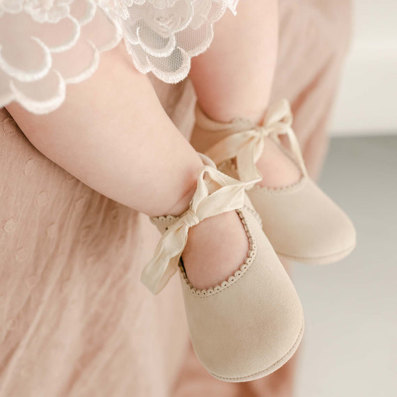 Close-up of a baby's feet in tan suede tie Mary Janes, held by a parent wearing a lace dress. The image conveys a gentle and affectionate moment.