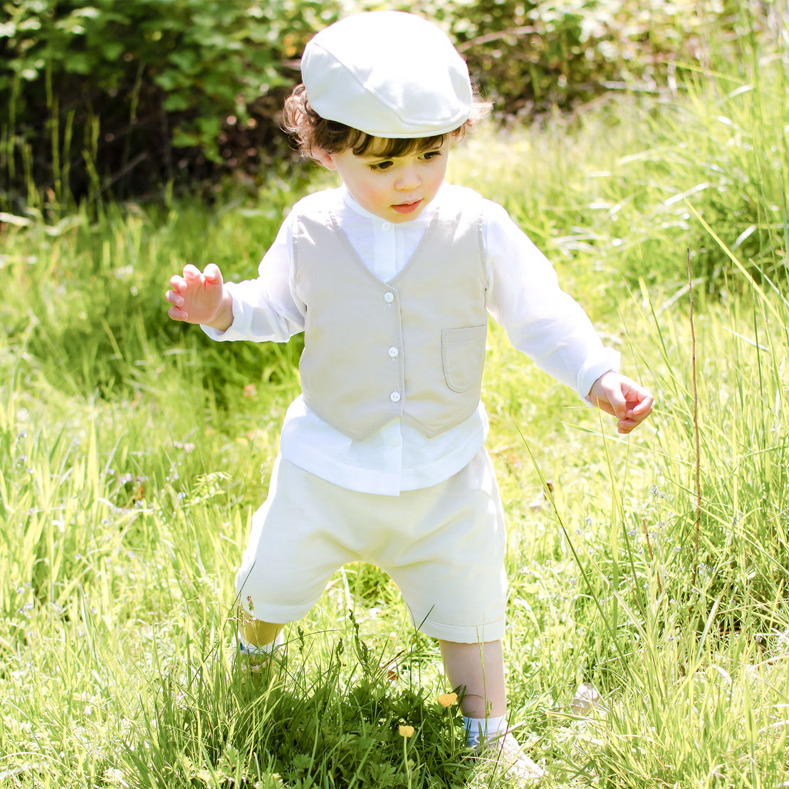 Photo of a baby boy outside in the forest. He is wearing the Silas Vest Suit that includes the vest, shorts, and onesie. He is also wearing the matching Silas newsboy cap