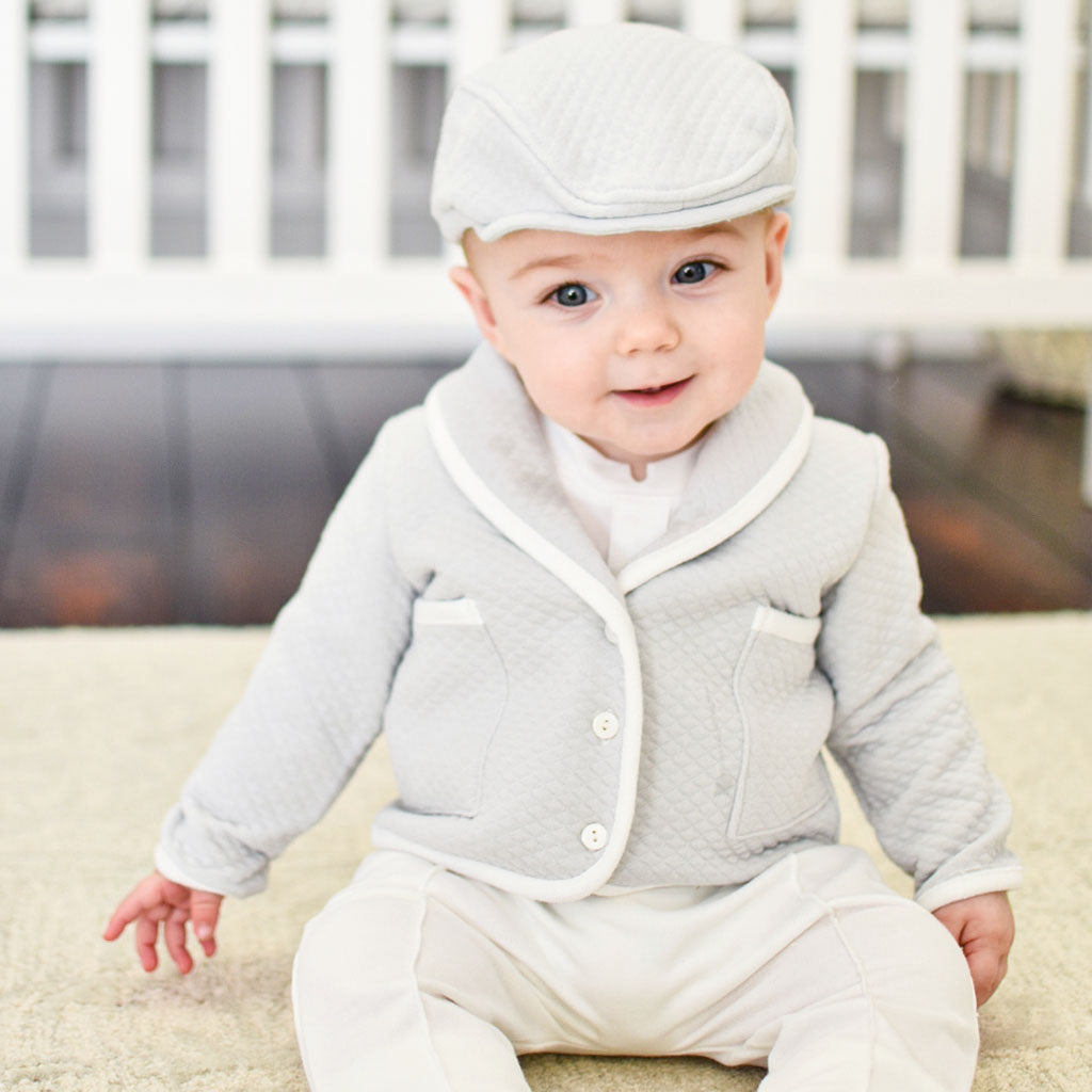 Baby boy sitting on ground smiling in his formal baby boy outfit, the Grayson 3-piece suit and newsboy cap.