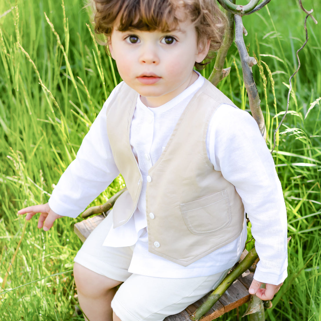 Photo of a baby boy sitting on a wooden chair outside in the forest. He is wearing the Silas Vest Suit that includes the vest, shorts, and onesie
