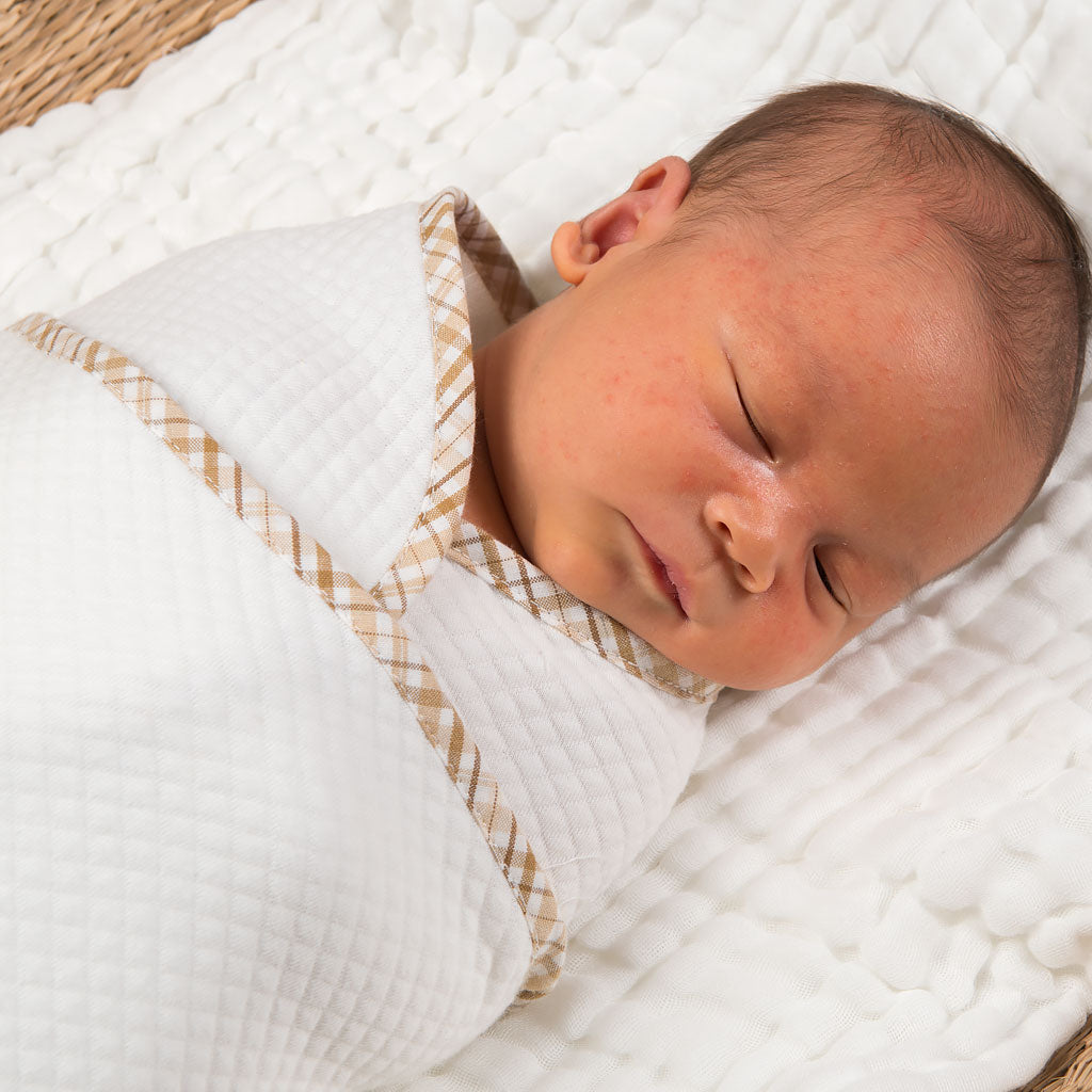A newborn baby sleeps peacefully, swaddled in a Dylan Personalized Blanket with a light brown trim, resting on a woven mat.