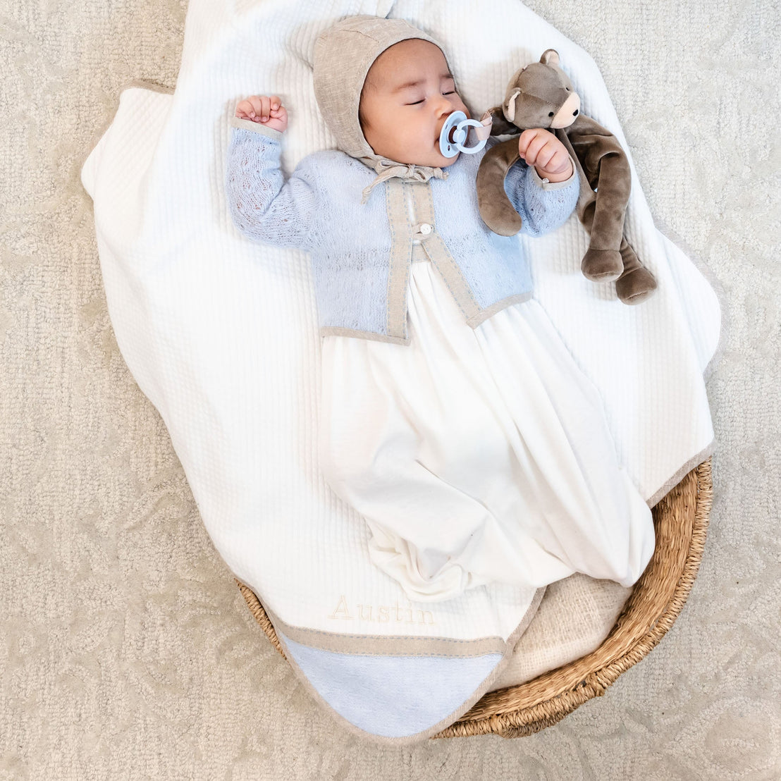 A newborn dressed in an Austin Layette & Hat, heirloom blue and white outfit with a bonnet, lies in a woven basket holding a plush bear. The baby has a pacifier and is on a cozy white blanket.