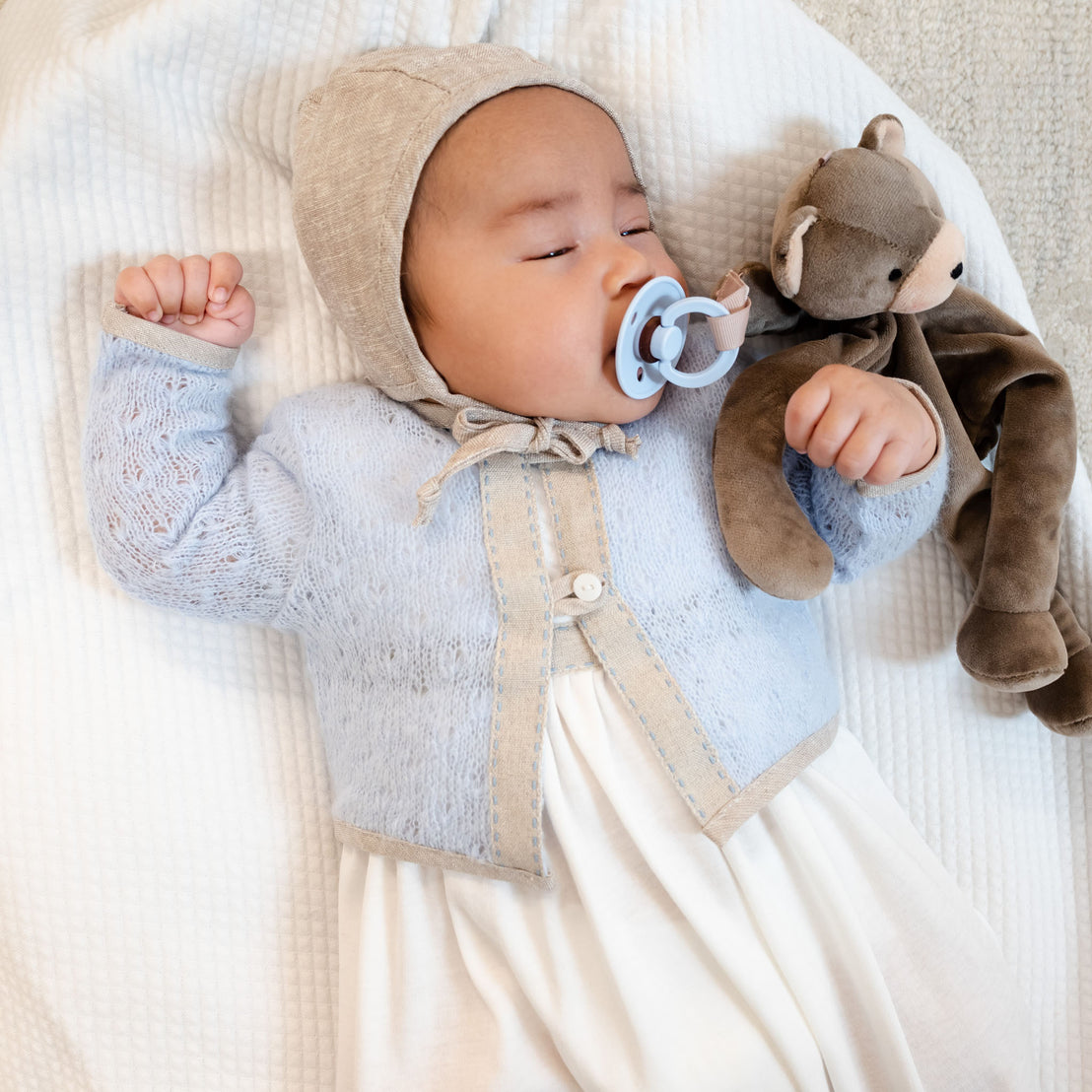 A baby wearing an Austin Sweater lies asleep on a white blanket, holding a plush bear and a pacifier in its mouth.
