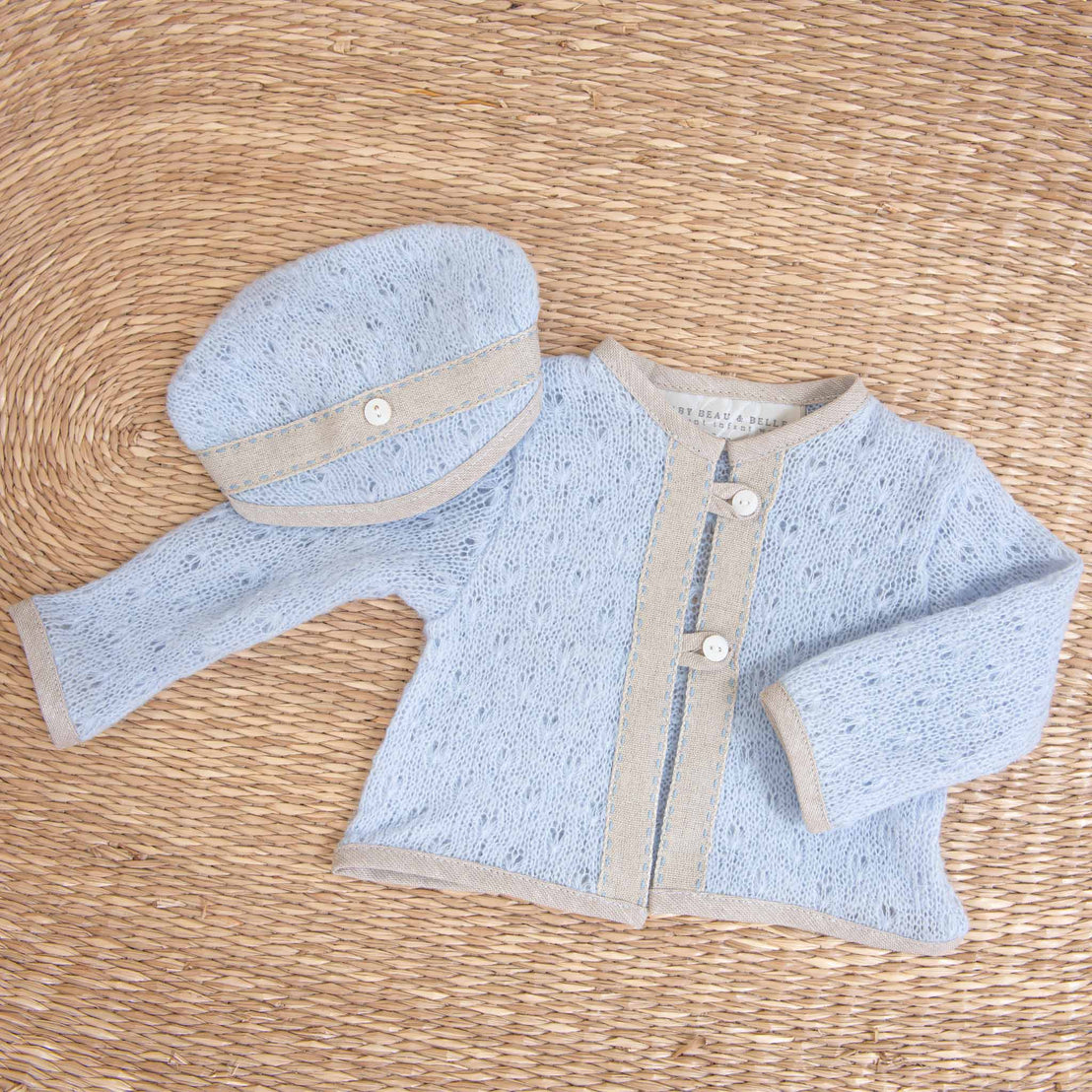 A light blue knitted Austin Newborn Knit Sweater and matching cap laid flat on a woven straw background, perfect for a coming home outfit.