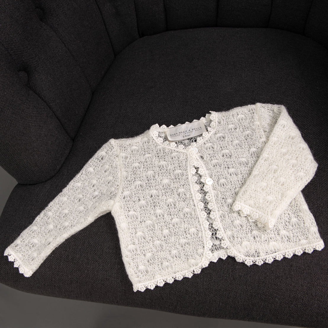 A delicate, hand-knit Grace Knit Sweater is neatly laid out on a dark gray, tufted chair. The sweater features intricate cotton lace patterns and a petite button closure.
