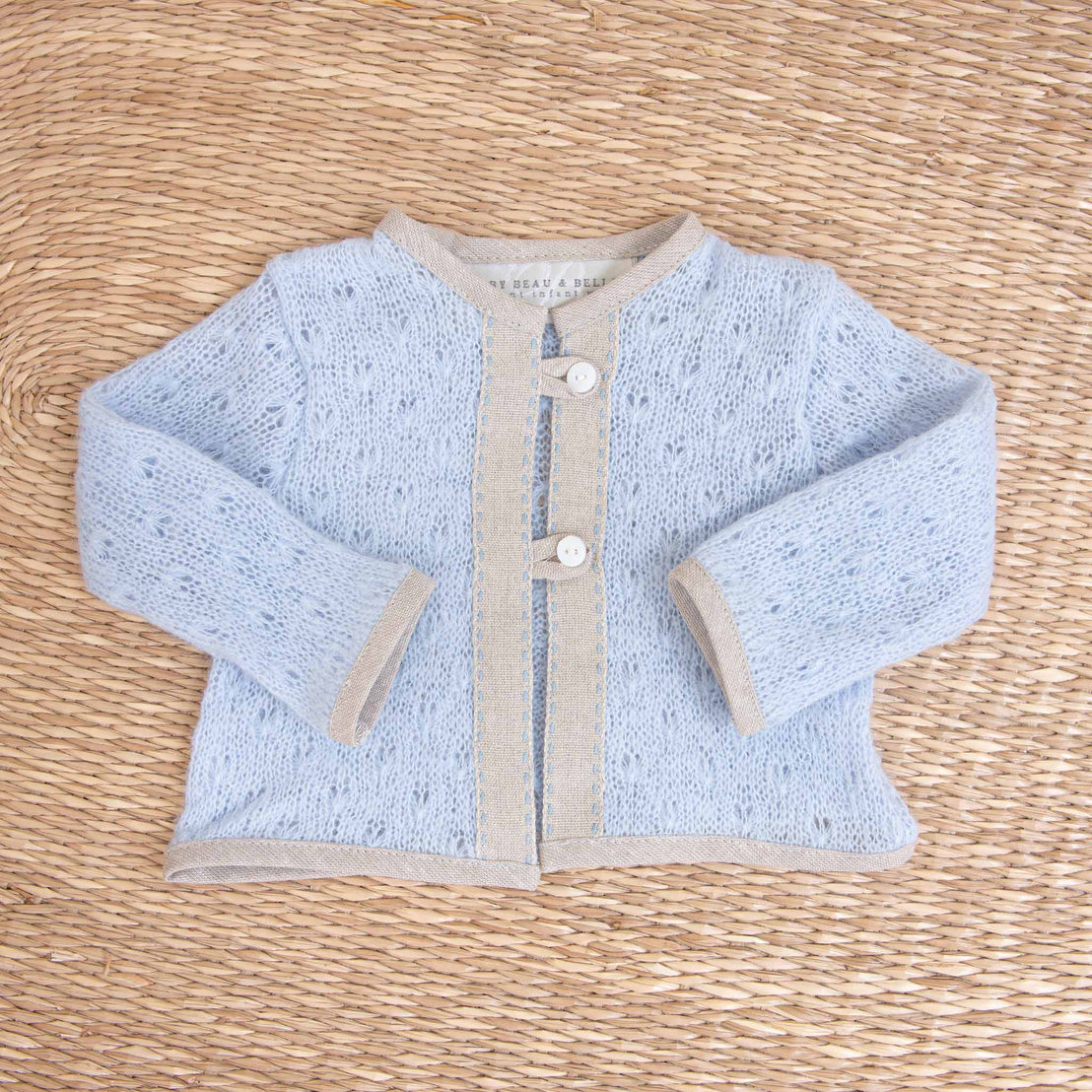 A baby's upscale, light blue Austin Sweater with white buttons lies flat on a woven straw background, featuring boutique beige knitted trim detailing.