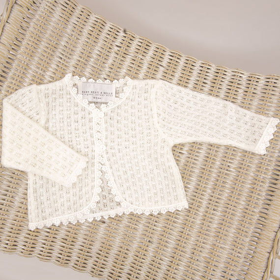 A delicate Eliza Knit Sweater laid flat on a woven wicker background, featuring intricate heirloom lace details and a front button closure.