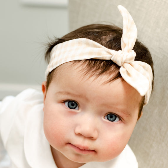 A close-up portrait of a newborn with big blue eyes and an Isla Tie Headband, looking directly at the camera with a soft, curious expression.