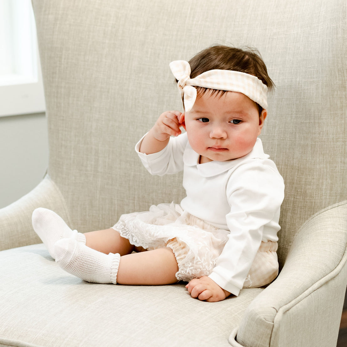 A baby girl wearing the Isla Bubble Skirt Set sits on a beige chair, looking thoughtful while touching her matching Isla Tie Headband.