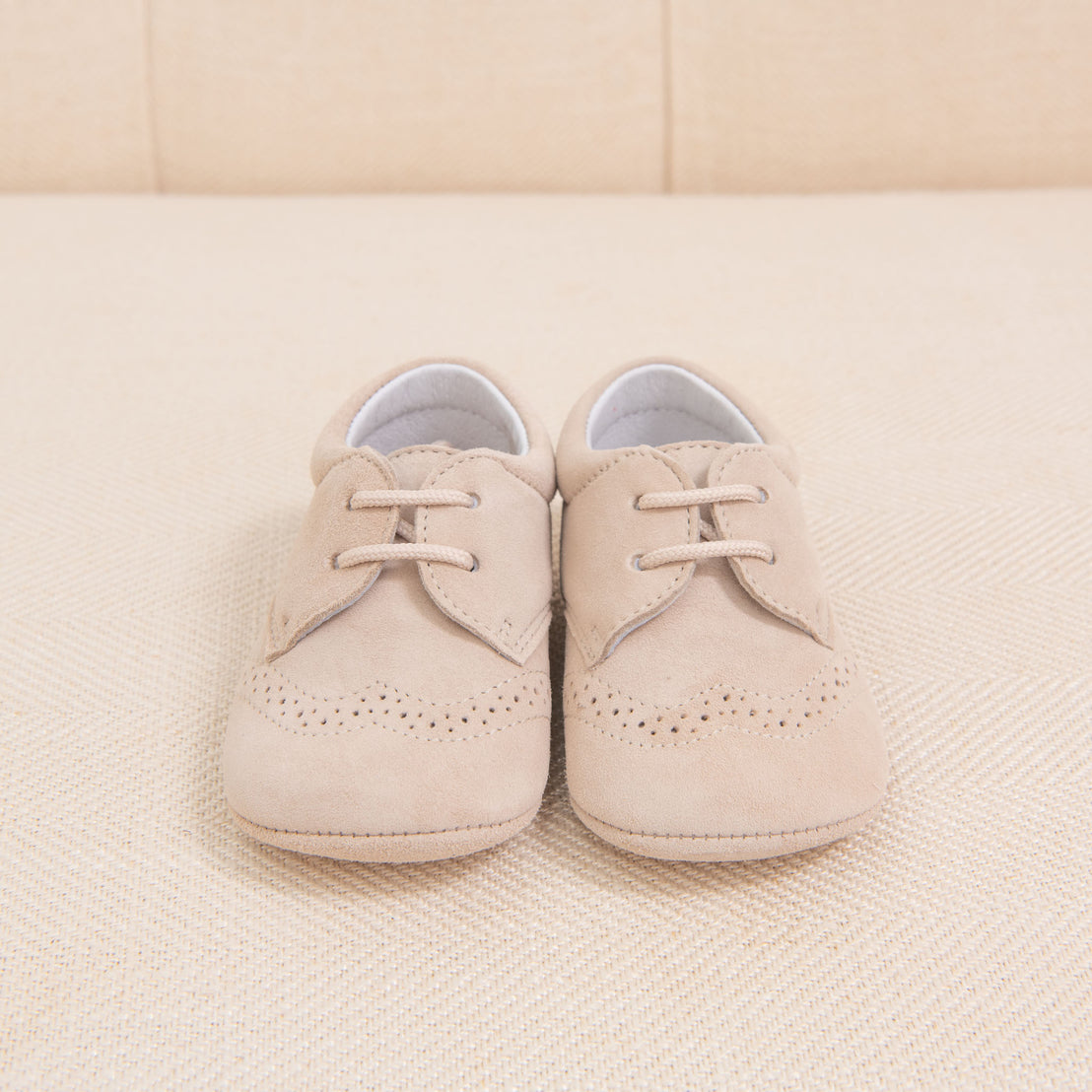 A pair of Baby Beau & Belle beige suede boys' shoes with laces and brogue detailing, placed neatly on a soft, textured beige background.