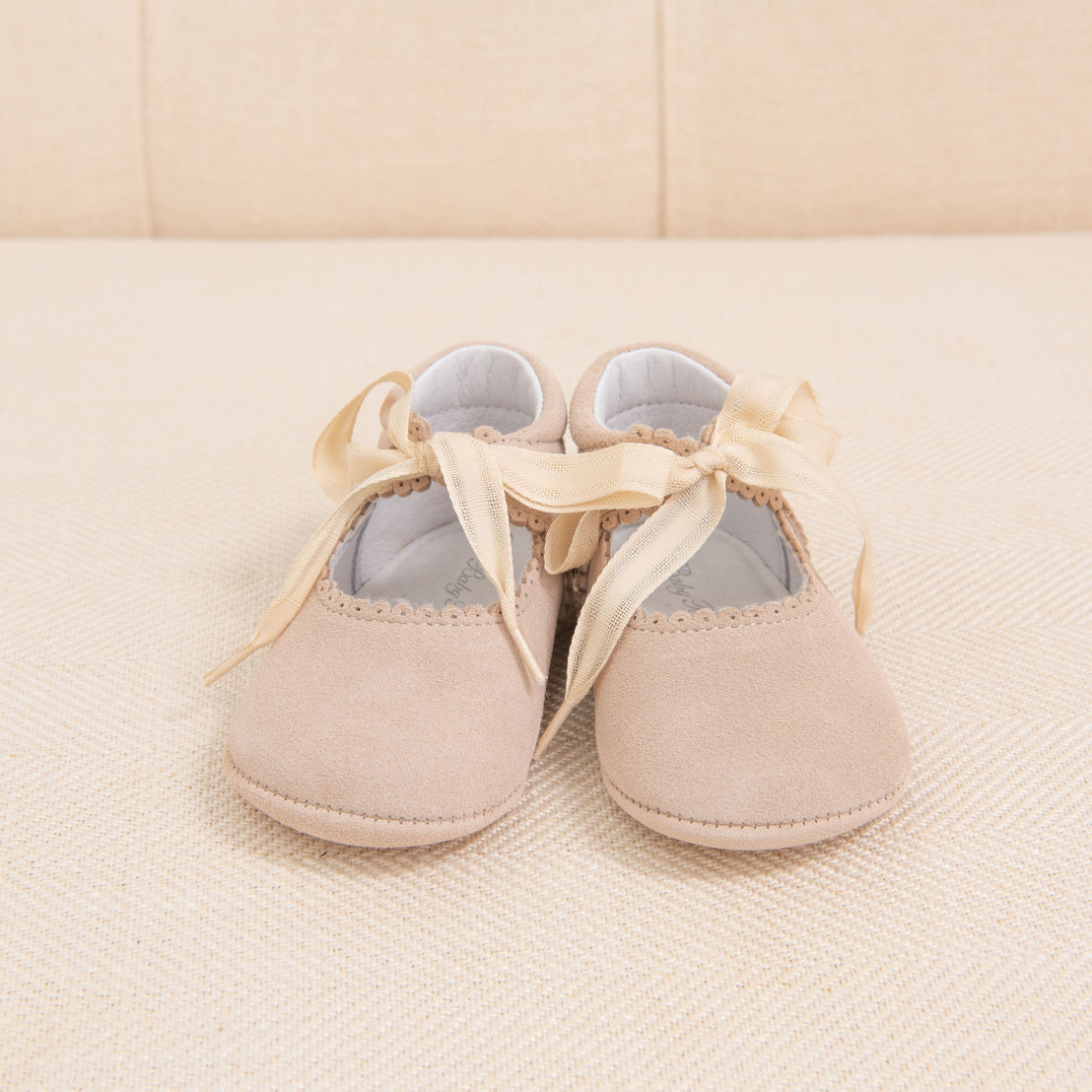 A pair of Girls Suede Tie Mary Janes in soft beige with delicate ribbon ties and scalloped edges, placed on a textured beige sofa.
