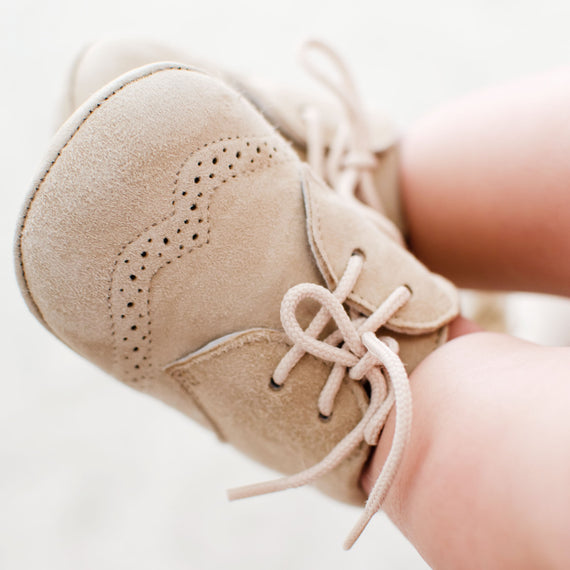 Close-up of a baby's foot in a Camel Suede Shoe with delicate lace-up detail and perforations on the side.