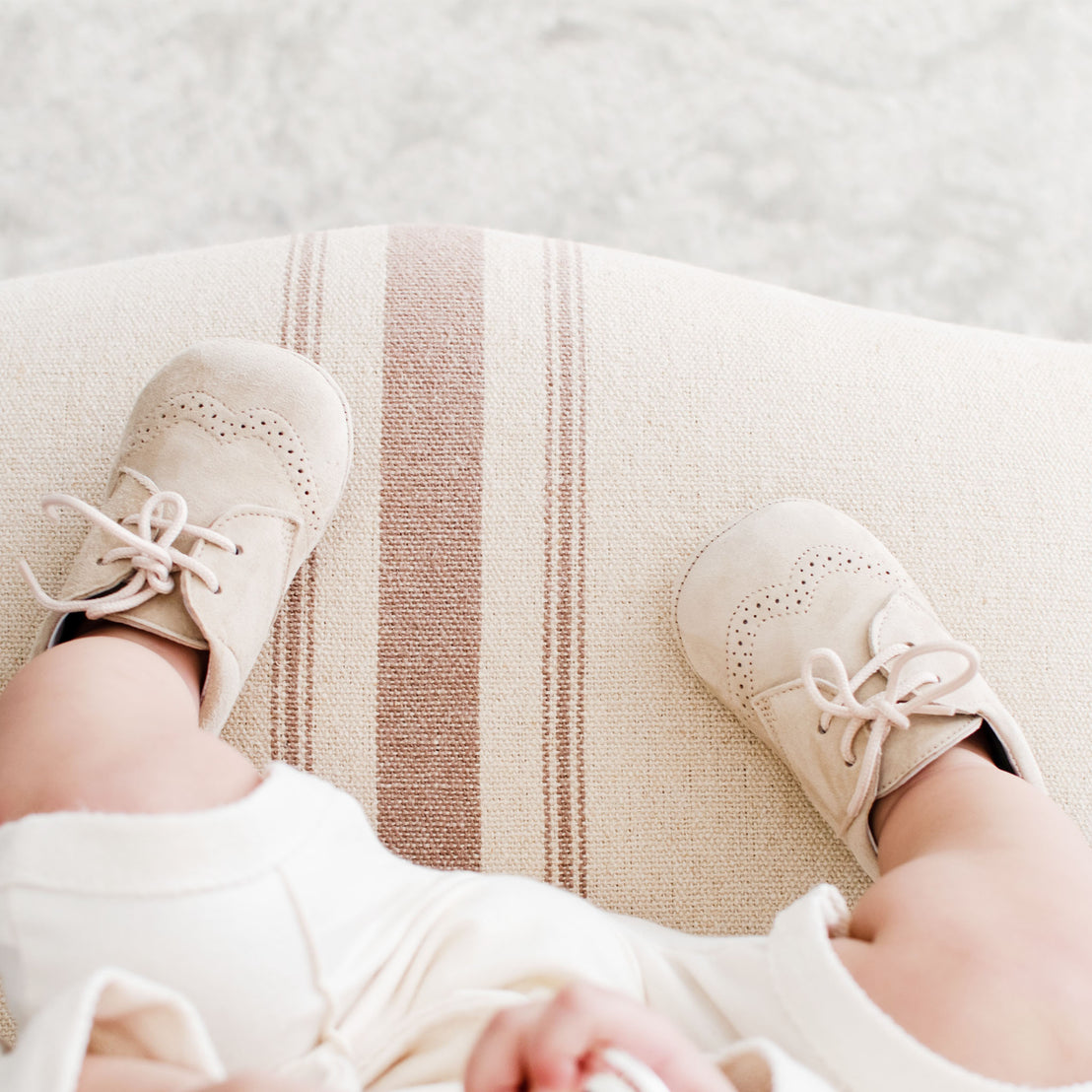 Photo of a baby wearing the Tan Suede Shoes on a chair. They are made with tan suede leather and feature detailed edging