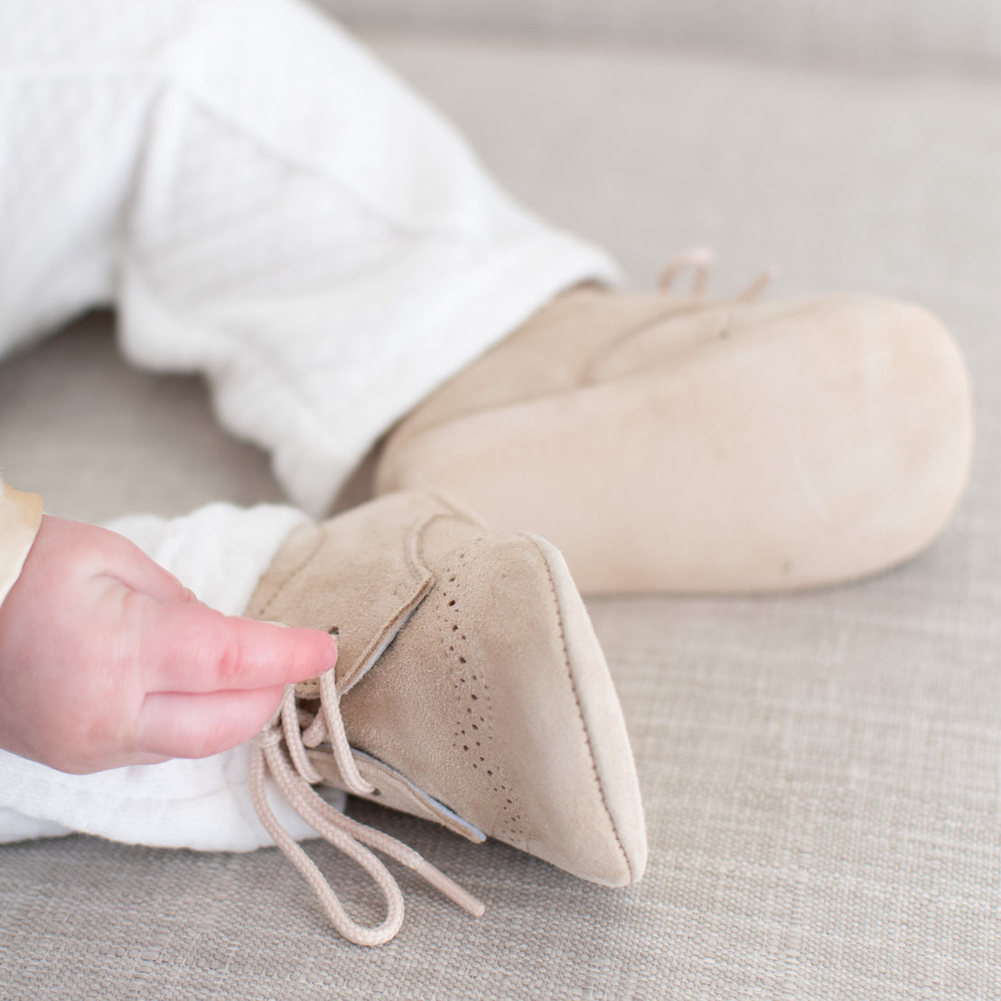 A close-up of a baby's feet in Camel Suede Shoes with laces, focusing on one hand touching the shoe. The background features a neutral soft fabric.