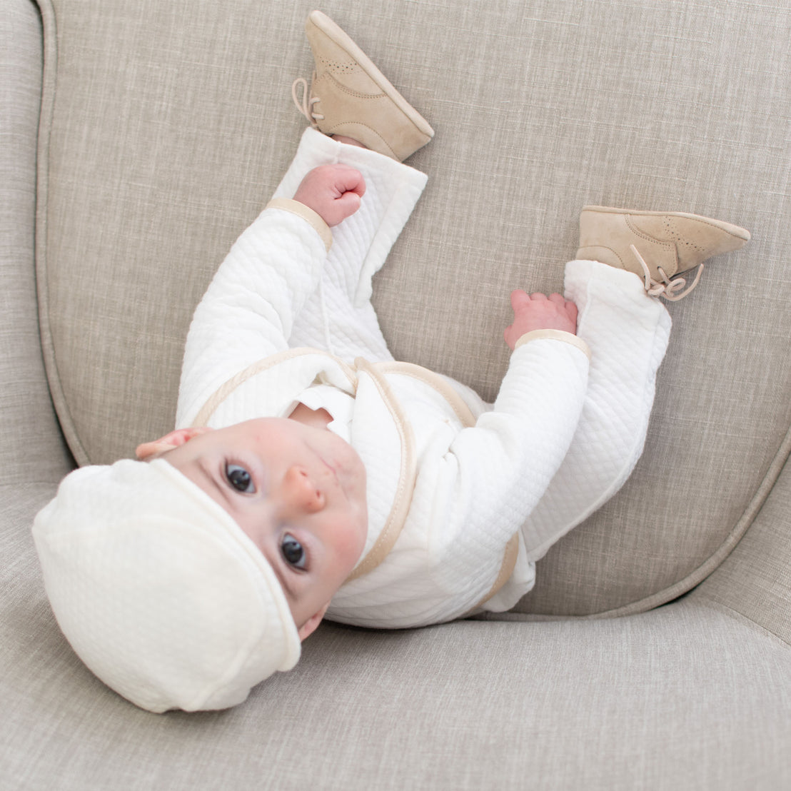 A baby dressed in a white outfit and hat lies on a gray sofa, looking up with wide blue eyes, wearing Camel Suede Shoes on its feet.