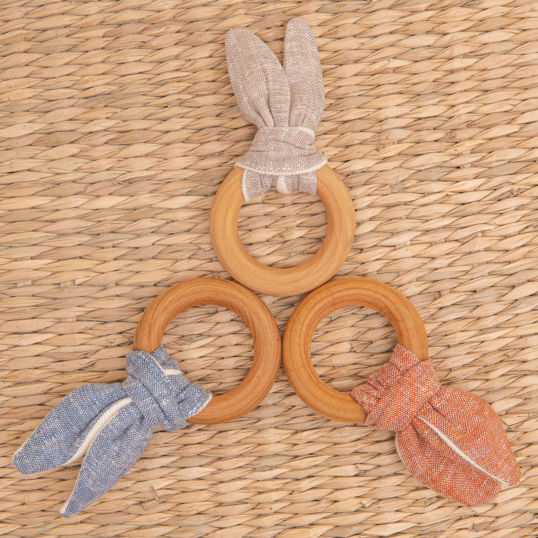  Flat lay photo of all three Silas Wooden Teether Rings, including the colors red, tan, and blue. The teether ring is made from organic maple wood and features a linen and french terry tie