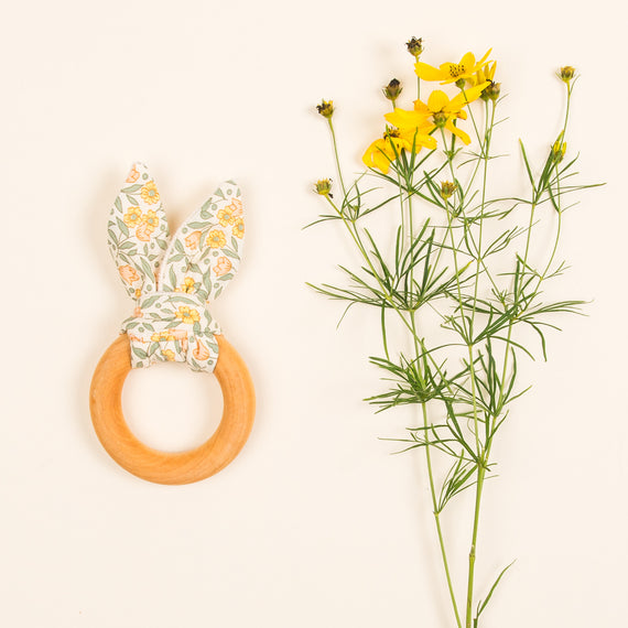 A Petite Fleur Wooden Teether Ring with fabric bunny ears next to a sprig of yellow flowers against a white background.