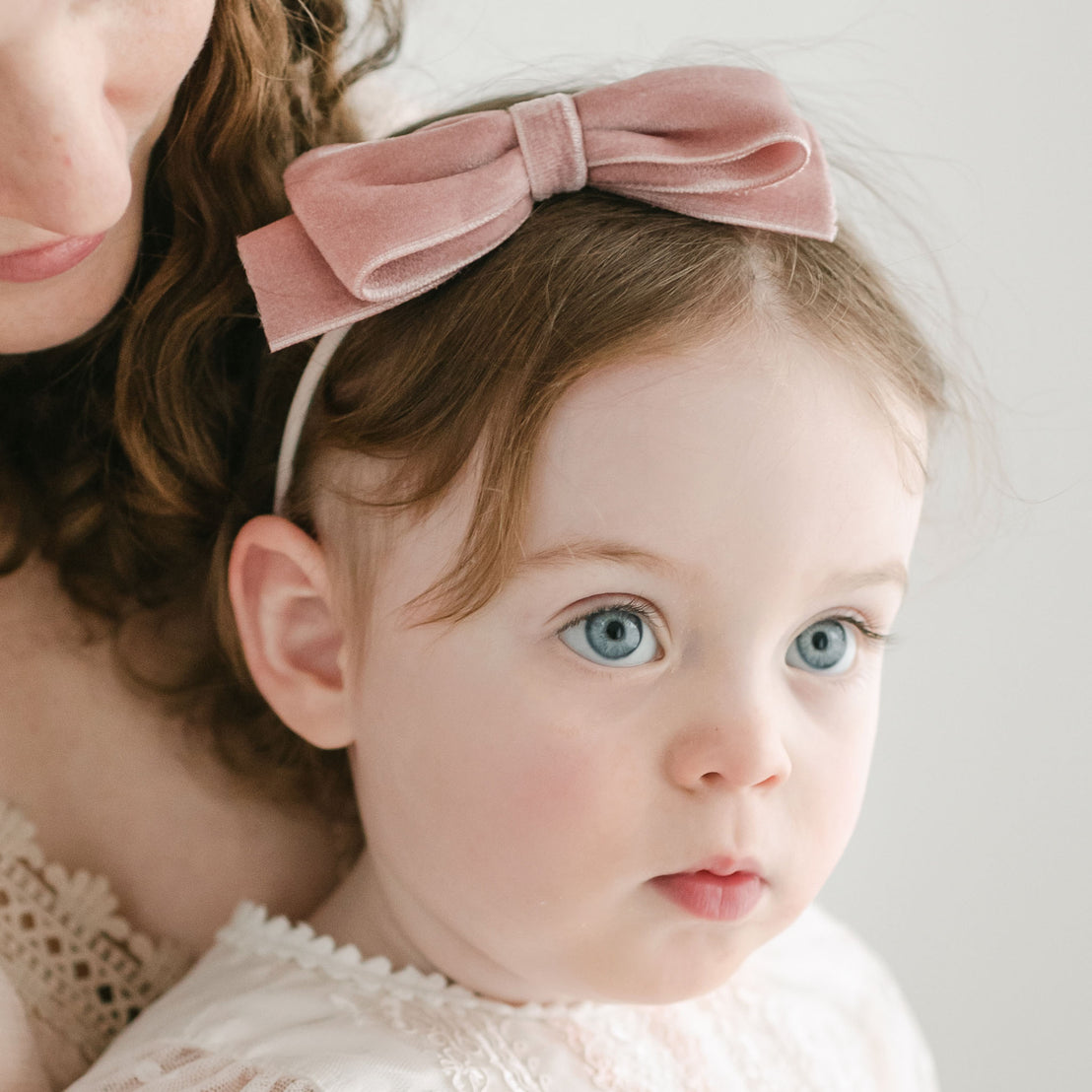 A close-up portrait of a baby with bright blue eyes wearing an Elizabeth Velvet Bow Headband in vintage pink, held by her mother whose face is partially visible as she embraces the child.