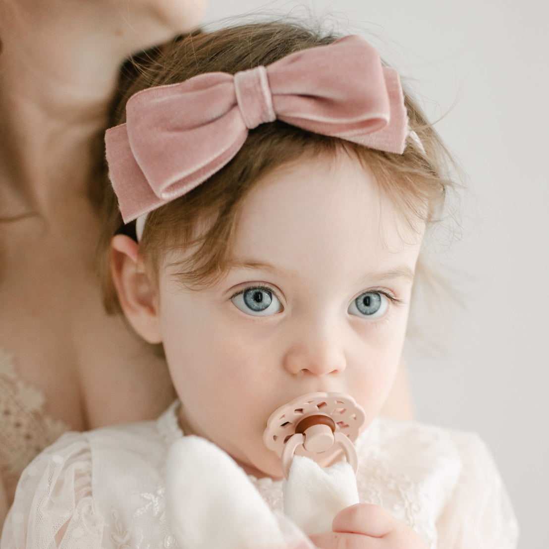 A close-up of a baby with big blue eyes, wearing a pink bow headband and a white lace Elizabeth Pacifier Set | Blush & Ivory outfit, being held by an adult (partially visible).