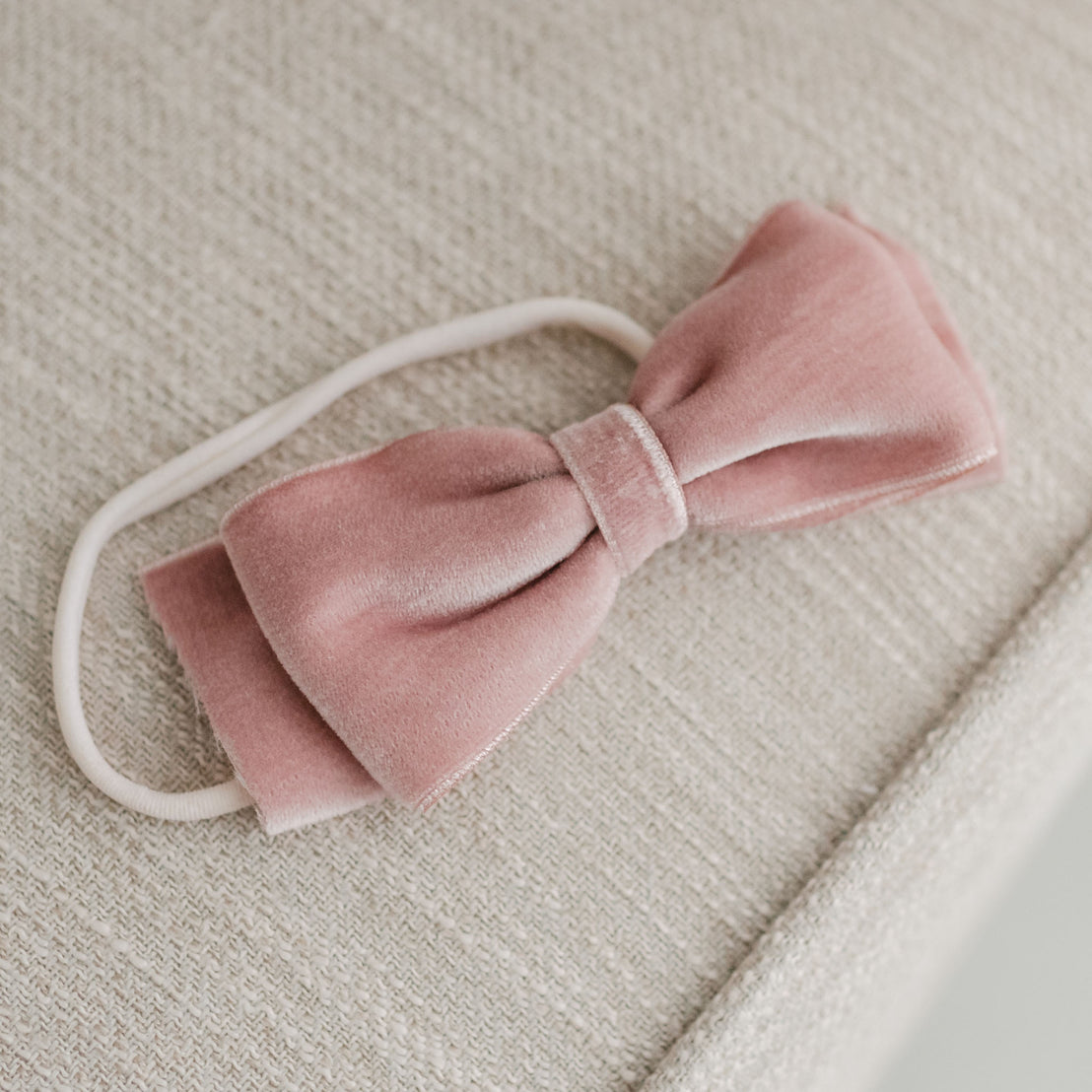 A soft pink Elizabeth Velvet Bow Headband with an elastic band lies on a textured beige fabric surface, suggesting a delicate and vintage piece.
