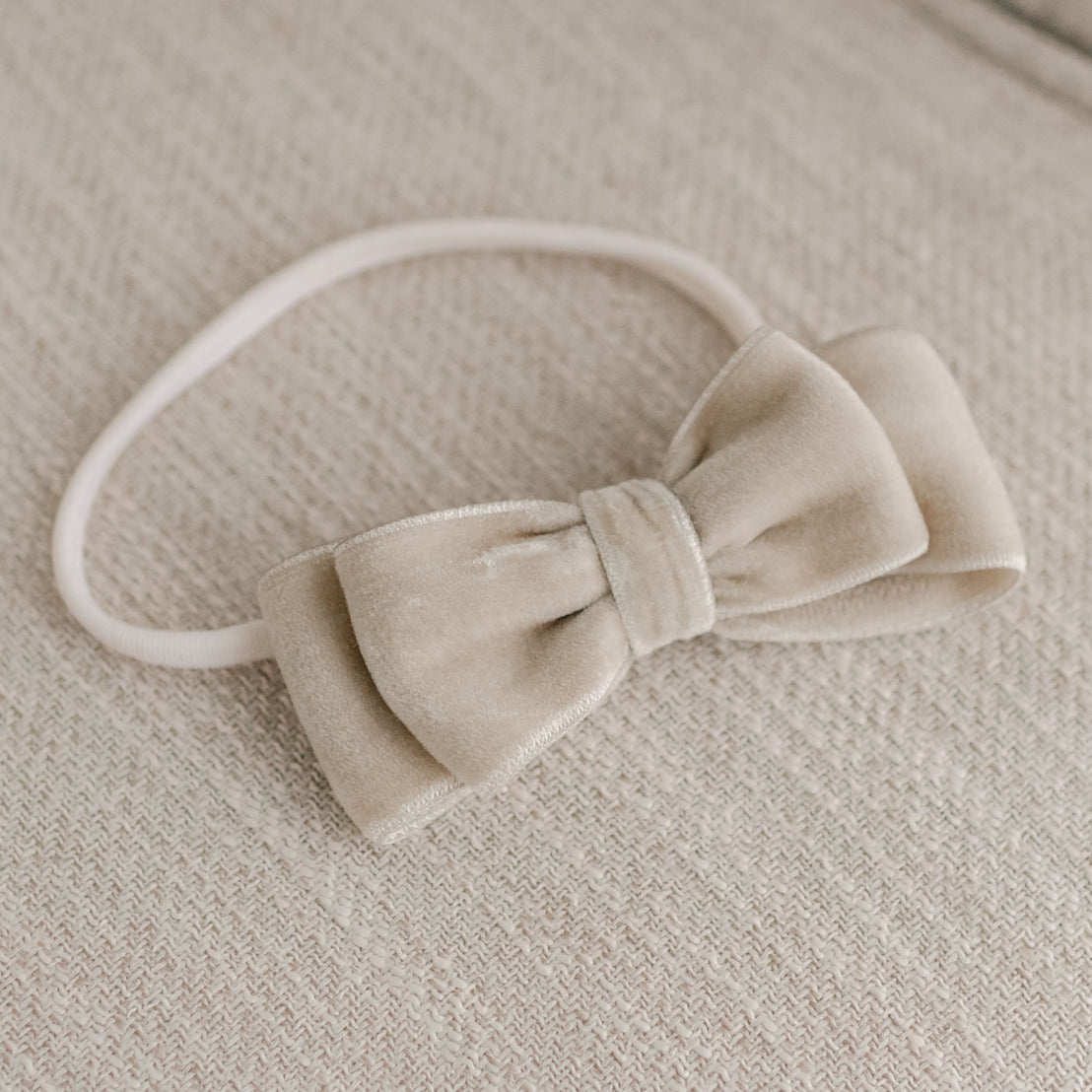 A beige Kristina Velvet Bow Headband attached to a matching elastic band, resting on a textured light grey fabric background, ideal for baptism or christening celebrations.