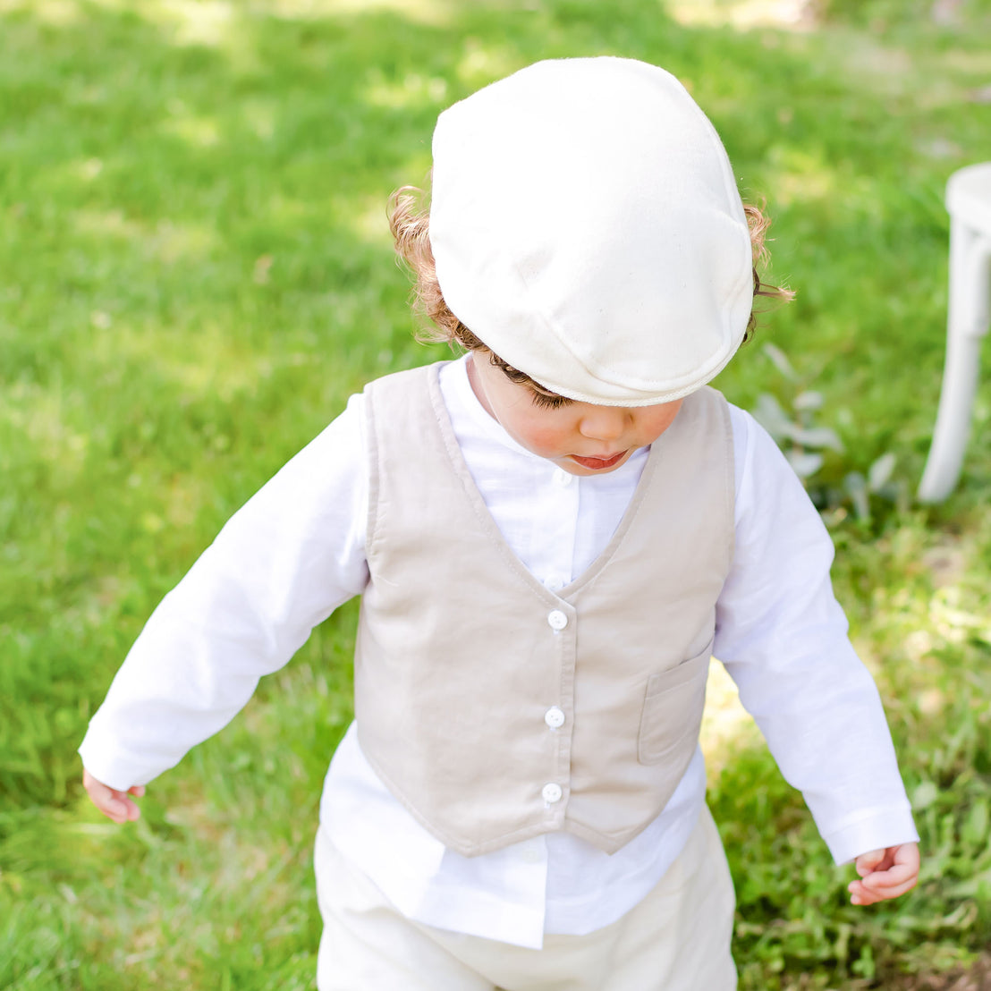 Photo of a baby boy outside in the forest. He is wearing the Silas Vest Suit that includes the vest, shorts, and onesie. He is also wearing the matching Silas newsboy cap
