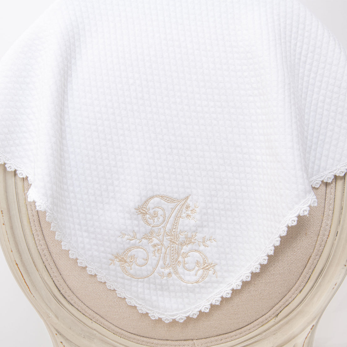 A white embroidered Initial Blanket with a detailed gold initial displayed on an elegant wooden chair.