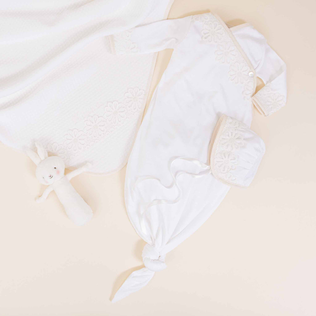 A flat lay of the Hannah Newborn Gift Set- Save 10%, featuring a white knitted gown with floral details, a matching hat, a cozy blanket, and a small white bunny plush toy, all arranged on.