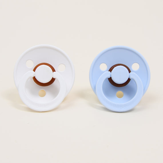 Two pacifiers side by side, the one on the left in white, the one on the right in baby blue. Both pacifiers are part of the Harrison Pacifier Set.