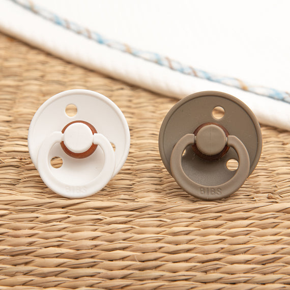 Two Mason Pacifier Sets, one white and one dark oak, lying on a textured woven mat.