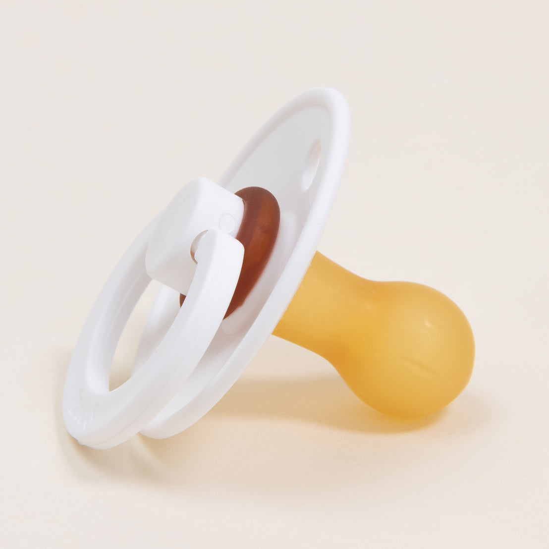 A close-up of a Bibs Pacifier with a white plastic ring and a yellow nipple on a light beige background.