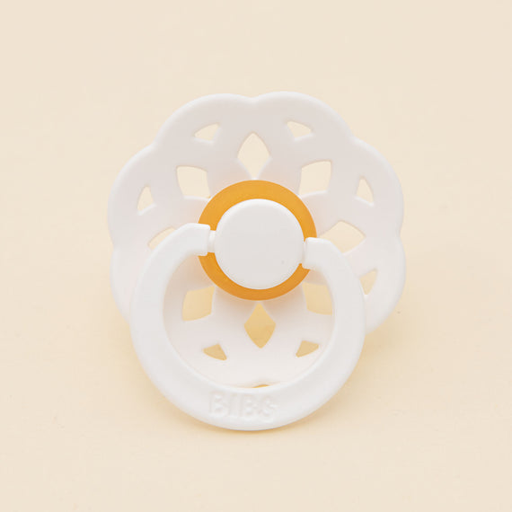 A Bibs Lace Pacifier in White with a hollow circular handle and petal-shaped shield, placed on a light beige background.